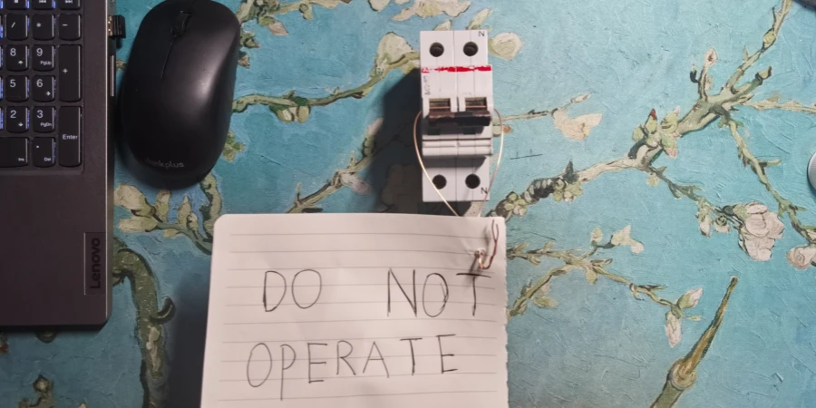 A circuit breaker secured with a red LOTO lock and white wire, with a handwritten DO NOT OPERATE note on lined paper, next to a Lenovo laptop and mouse on a blue floral surface.