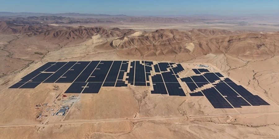 An aerial view of the Leghrous 220 MWp solar plant in Biskra Province, Algeria, showing rows of dark solar panels on sandy desert terrain surrounded by rocky hills and distant mountains.