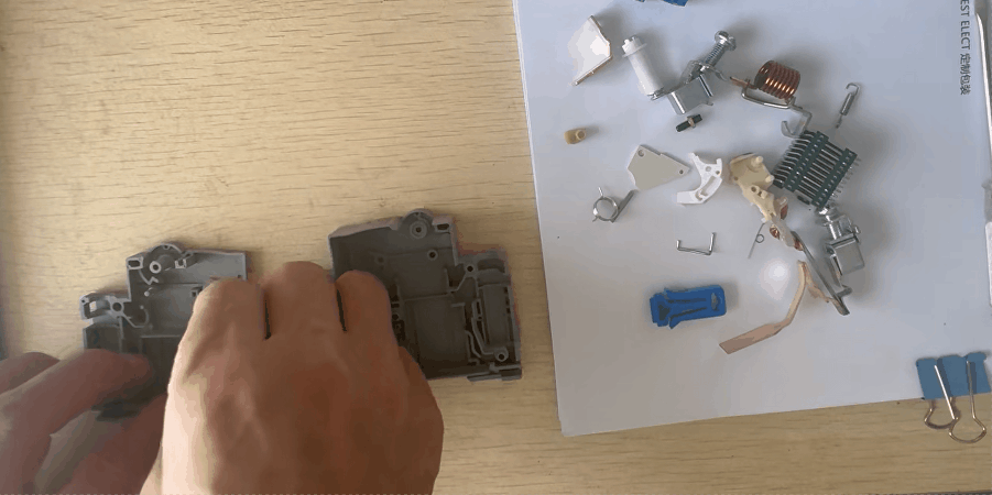 A short animated GIF clip showing a person's hands assembling a small circuit breaker on a wooden table. Two gray plastic housings are held in the hands, while various small internal components such as springs, coils, metal plates, and a blue plastic piece are laid out on a white surface to the right.