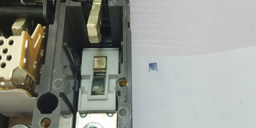 A close-up of an MCCB with its cover removed, showing the internal contacts and a white paper with carbon paper beside it, marked with a small blue square.