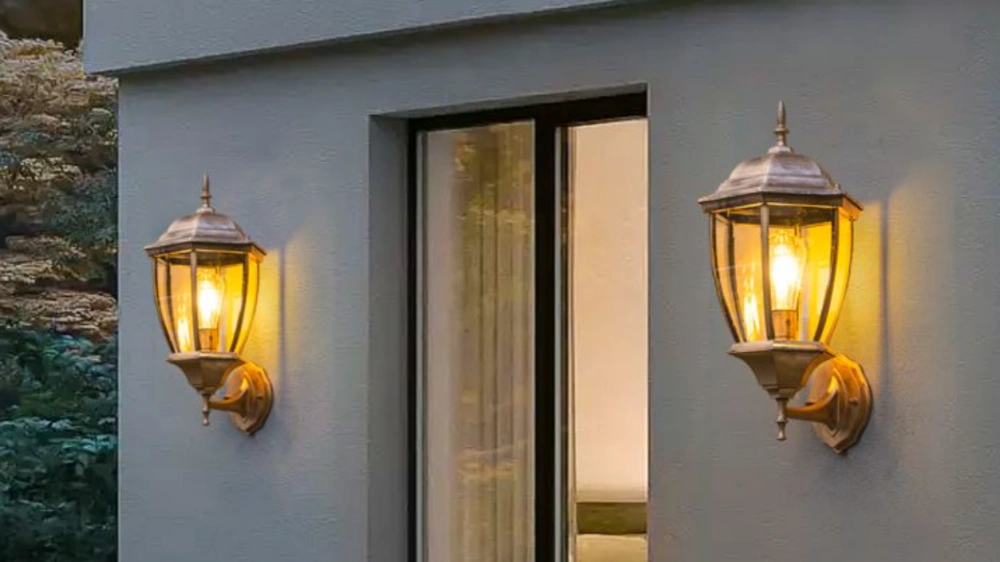 Two illuminated brass outdoor wall lanterns mounted on a gray wall, flanking a large glass window with sheer curtains.
