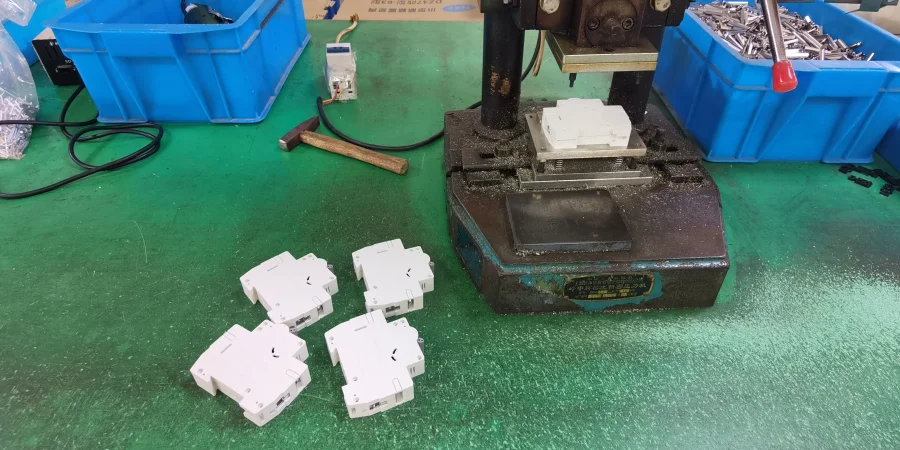 A riveting machine on a green workbench with white miniature circuit breaker housings placed on its base and nearby, ready for pressing.
