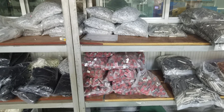 Two wooden shelves holding clear plastic bags filled with various miniature circuit breaker components, including dark metal parts, red and silver pieces, and loose hardware, in a storage area with a window in the background.