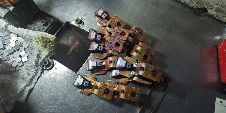 A pile of copper static contacts with welded square silver tips, resting on a metal work surface on an MCCB production line, next to a pile of loose silver contact pieces and a cloth