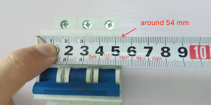 A hand holds a tape measure across a 3P miniature circuit breaker, showing a width of about 54 mm with a red arrow and text annotation.