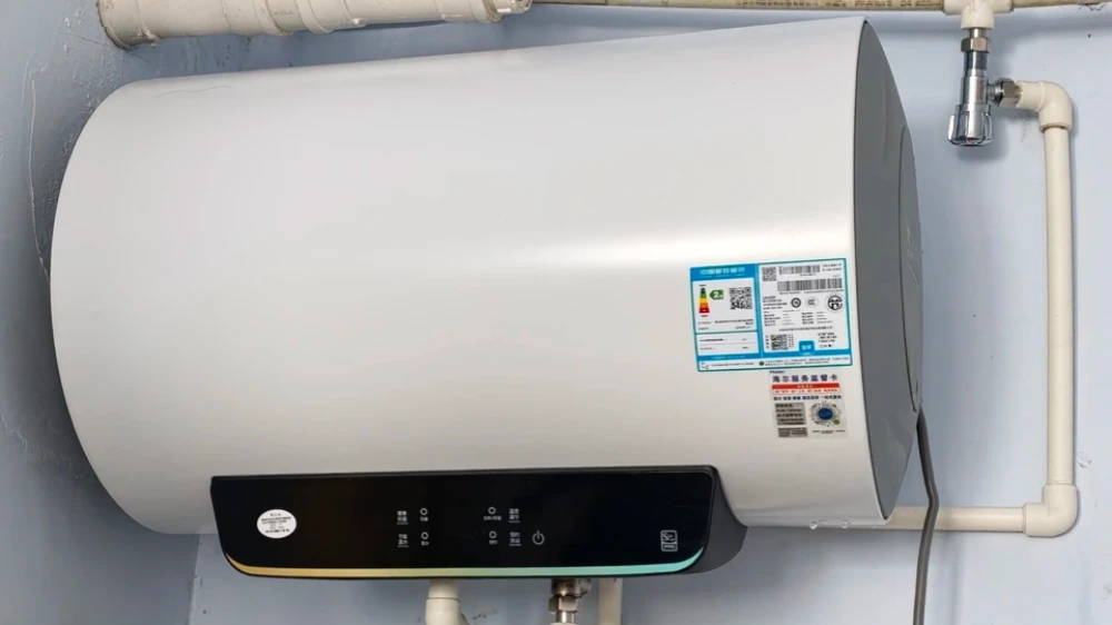 A white cylindrical electric water heater mounted on a wall, with a black digital control panel at the bottom and white pipes connecting to it.