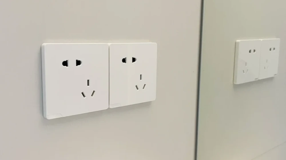 Two white electrical wall sockets mounted side by side on a light gray wall, with their reflection visible on a mirrored surface to the right.
