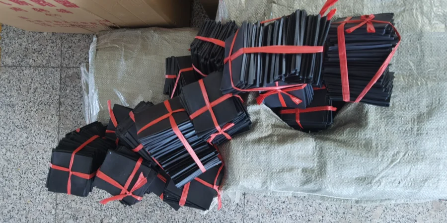Several stacks of flat black phase barriers for molded case circuit breakers, bundled with red plastic straps on a woven sack over a speckled floor, with a cardboard box in the top left corner