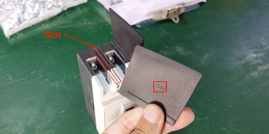 A hand holding a black interphase barrier above a circuit breaker, with a red arrow pointing to a slot labeled 'Slot' and a red box highlighting a small notch on the barrier.
