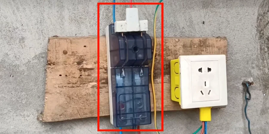 A knife switch is mounted on a wooden board against a concrete wall, with a wall socket beside it.