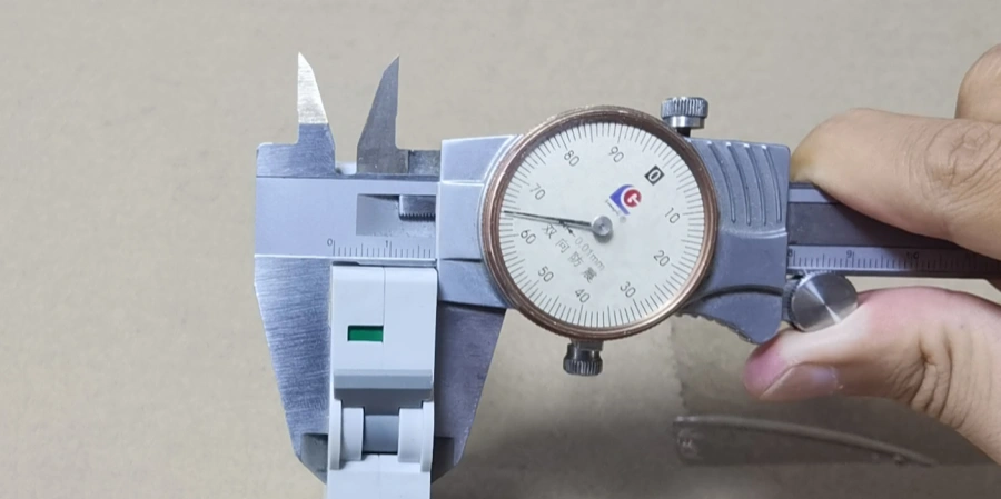A hand holds a dial caliper measuring the width of a white mcb