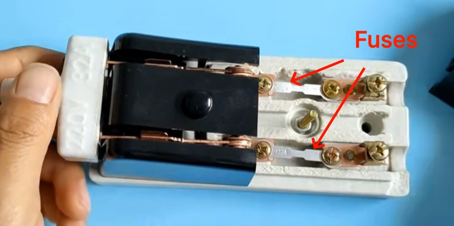 A hand holding an open white knife switch against a blue background. A red arrow labeled Fuses points to two glass fuses inside.