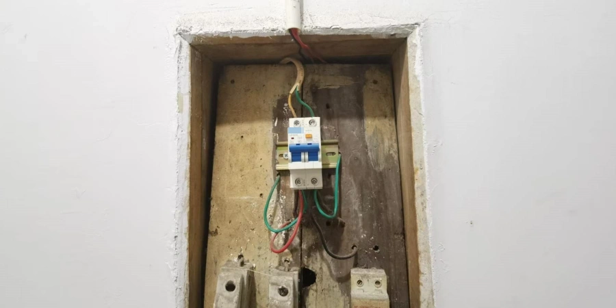 A white RCBO mounted on a wooden board inside a wall recess, with green and red wires connected to it.