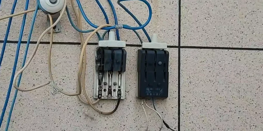 Two surface-mounted knife switches on a speckled tiled wall, with exposed wires and blue cables running around them.