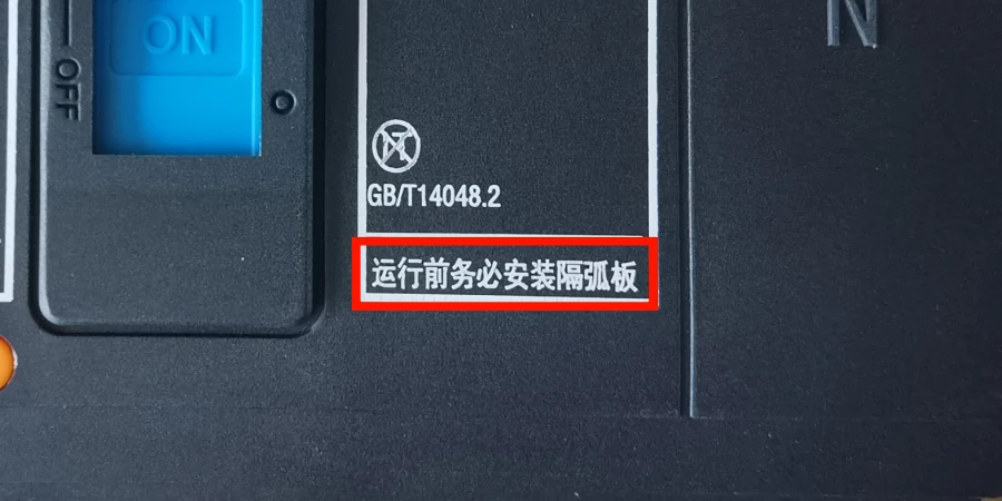 Close-up of a circuit breaker panel showing a blue ON/OFF switch, the standard number GB/T14048.2, and a red-outlined warning label with Chinese text meaning 'Install the interphase barrier before operation'.