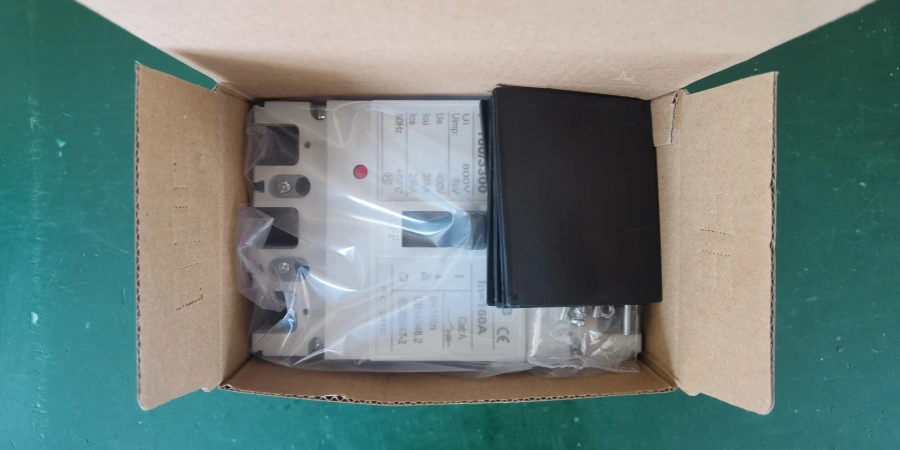 An mccb wrapped in plastic next to black interphase barriers inside an open cardboard box on a green surface